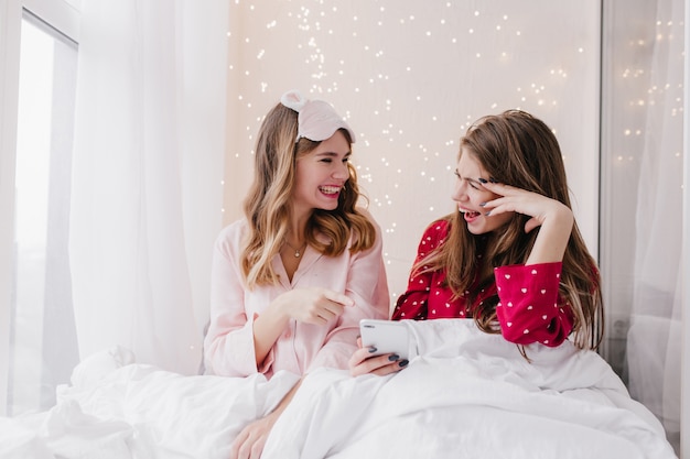 Splendide ragazze bianche che scherzano mentre posano la mattina presto. Foto interna di sorelle emotive in pigiama carino che si guardano con un sorriso.