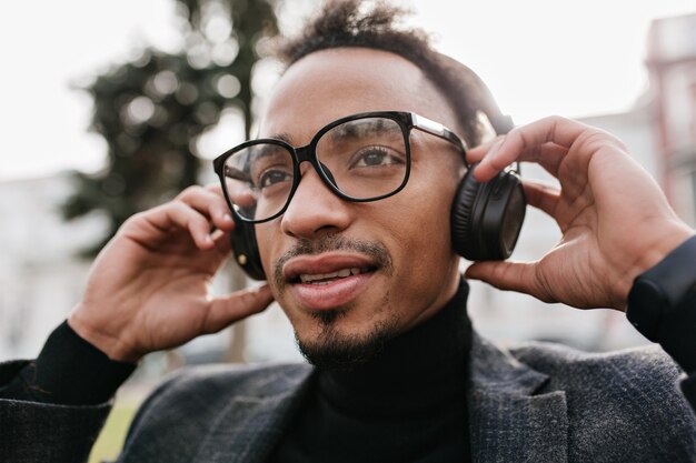 Splendido mulatto dagli occhi scuri che tocca le sue cuffie. Ritratto del primo piano del ragazzo africano castana fiducioso in vestito che ascolta la musica al mattino.