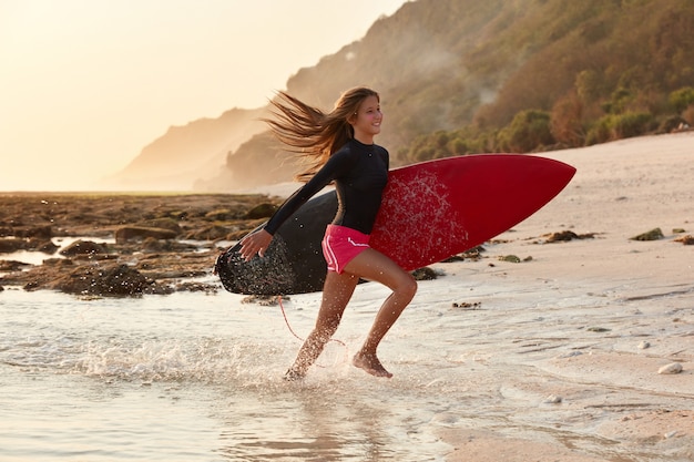 Sport acquatici e concetto di tempo di ricreazione. Il cavaliere felice dell'onda corre sulla spiaggia, vestito in costume da bagno, ha un'espressione positiva