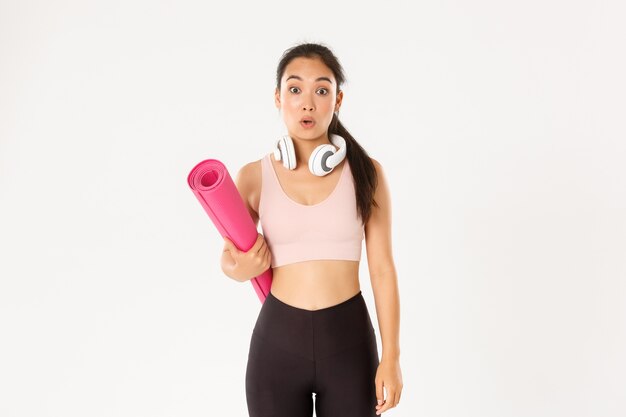 Sport, benessere e concetto di stile di vita attivo. Ragazza asiatica confusa e scioccata fitness, non riesco a capire perché la palestra si è chiusa, tenendo il tappetino di gomma e guardando sorpreso dalla fotocamera, sfondo bianco.