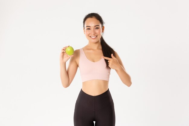 Sport, benessere e concetto di stile di vita attivo. Ritratto della ragazza asiatica sottile e in forma sorridente di forma fisica, consiglio dell'allenatore di allenamento che mangia le vitamine e l'alimento sano, indicante la mela verde, fondo bianco.
