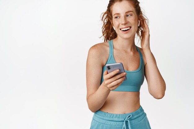 Sport e fitness Atleta femminile sorridente che ascolta musica nelle cuffie che tengono il telefono cellulare in piedi in abbigliamento sportivo su sfondo bianco