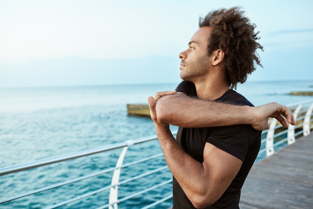 Sport, fitness e stile di vita sano. Corridore afroamericano in forma che sembra concentrato mentre allunga le braccia in riva al mare, facendo esercizi di allungamento delle braccia e delle spalle