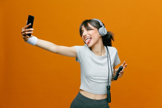 Sportiva bella donna in abbigliamento sportivo con le cuffie utilizzando smartphone facendo selfie felice e positivo sorridente allegramente sporgendo lingua in piedi su sfondo arancione