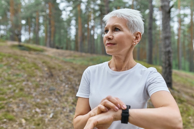 Sportiva di mezza età autodeterminata in maglietta bianca che regola orologio intelligente, controlla le statistiche di fitness, monitorando le sue prestazioni di corsa durante l'allenamento cardio nel parco