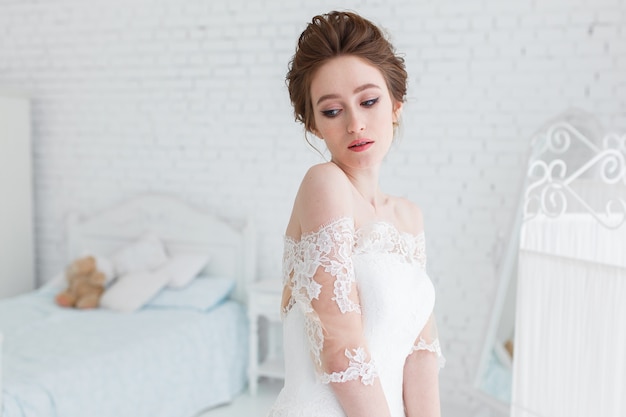 Sposa che posa in vestito da sposa nello studio su un fondo di un muro di mattoni e di un letto bianco