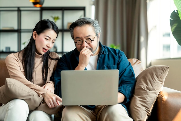 Sposare le finanze domestiche delle coppie asiatiche rivedendo i loro conti bancari utilizzando laptop e calcolatrice in soggiornocoppia asiatica seduta sul divano con laptop ed emozione stressante a casa
