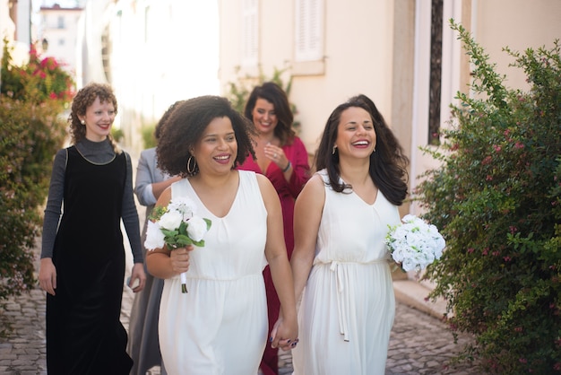 Spose felici e ospiti al matrimonio. Donne sorridenti con mazzi di fiori che si tengono per mano andando da qualche parte