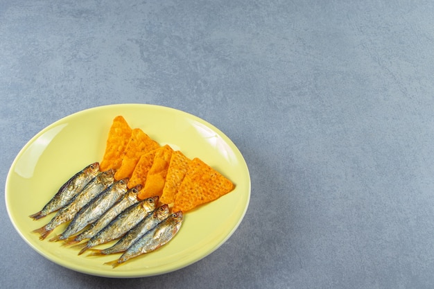 Spratti salati e patatine su un piatto accanto a un bicchiere di birra, crostini di pane e semi, sullo sfondo di marmo.