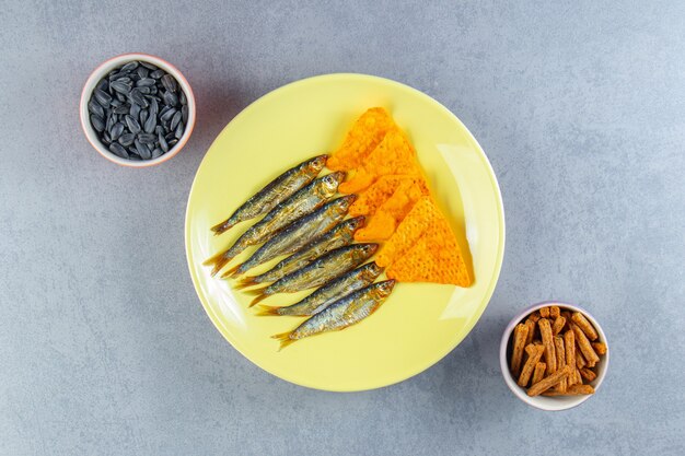 Spratti salati e patatine su un piatto accanto a un bicchiere di birra, crostini e semi, sulla superficie di marmo.