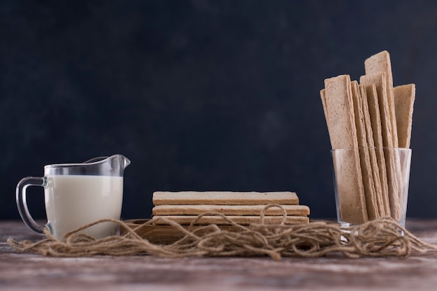 Spuntini e cracker nel piatto di legno con un bicchiere di latte su sfondo nero.