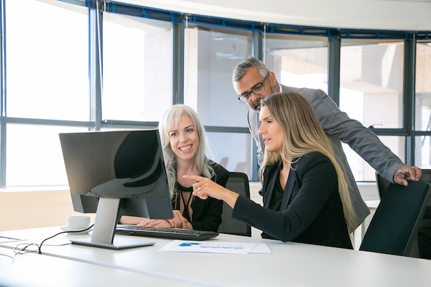 Squadra di affari di successo che guarda insieme il contenuto sul monitor del computer, discutendo del progetto, seduto al posto di lavoro e indicando il display. Comunicazione aziendale o concetto di lavoro di squadra