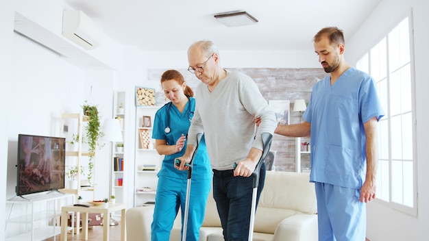 Squadra di infermieri o assistenti sociali che aiutano un vecchio disabile a camminare con le sue stampelle fuori dalla stanza della casa di cura.