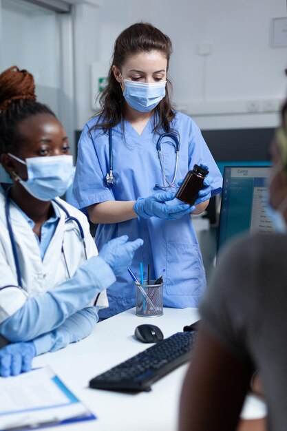 Squadra ospedaliera che discute con un paziente malato afroamericano che spiega il trattamento farmacologico durante la consultazione clinica in studio medico. Donne che indossano una maschera protettiva contro il coronavirus