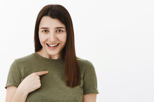 Stai parlando di me. Ritratto di affascinante bruna sorpresa e compiaciuta di 20 anni che indossa una maglietta verde oliva che sorride felicemente mentre indicava il suo petto interrogato, chiedendo se è stata scelta su un muro grigio
