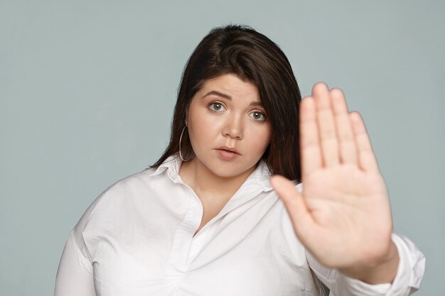 Stammi lontano. Segretaria femminile paffuta giovane bruna dispiaciuta seria in abbigliamento formale fissando, mano protesa, facendo il gesto di arresto con il palmo, esprimendo rifiuto o rifiuto