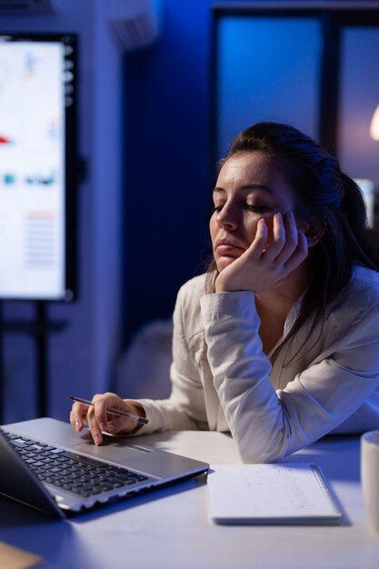 Stanca donna remota che parla pisolino sulla sedia che lavora al computer portatile che analizza le statistiche finanziarie