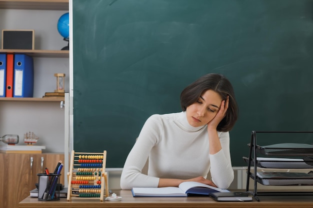 stanco con gli occhi chiusi che mettono la mano sulla testa giovane insegnante seduta alla scrivania con gli strumenti della scuola in classe