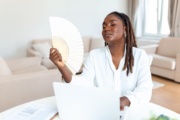 Stanco donna africana lavorare su laptop a casa ufficio onda con ventilatore a mano soffre di colpo di calore all'interno Malato esausto giovane uso femminile vacillare respirare aria fresca mancanza di condizionatore di ventilazione