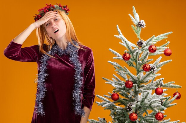 Stanco giovane bella ragazza in piedi vicino all'albero di Natale che indossa un abito rosso e la corona con la ghirlanda sul collo mettendo mano sulla fronte isolata su sfondo arancione