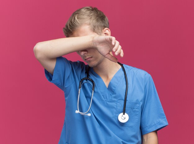 Stanco giovane medico maschio che indossa l'uniforme del medico con lo stetoscopio che pulisce la fronte con la mano isolata sulla parete rosa