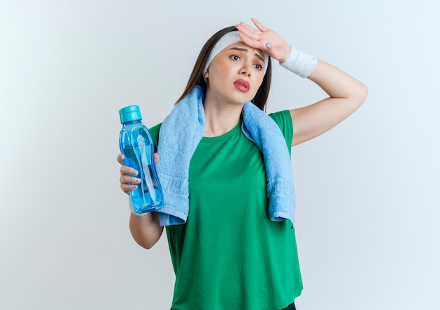 Stanco giovane sportivo da donna che indossa la fascia e braccialetti con asciugamano intorno al collo tenendo la bottiglia di acqua mettendo la mano sulla fronte guardando a lato