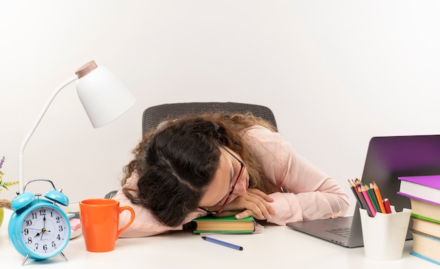 Stanco giovane studentessa graziosa con gli occhiali seduto e dormire alla scrivania con strumenti di scuola isolati su sfondo bianco