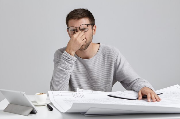 Stess, superlavoro e scadenza. Stanco giovane architetto con la barba lunga che massaggia il ponte nasale