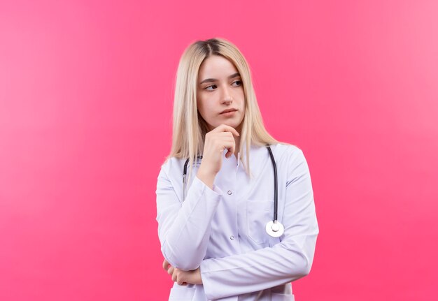 Stetoscopio d'uso della giovane ragazza bionda del medico di pensiero in abito medico mise la mano sul mento sulla parete rosa isolata