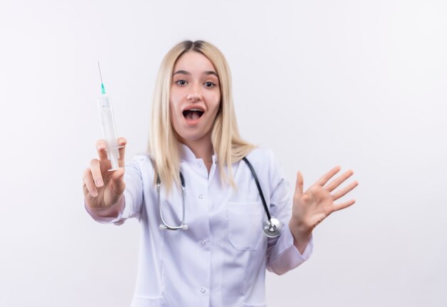 Stetoscopio da portare della giovane ragazza bionda del medico allegro e camice medico in parentesi graffa dentale che tiene fuori la siringa alla macchina fotografica sulla parete bianca isolata