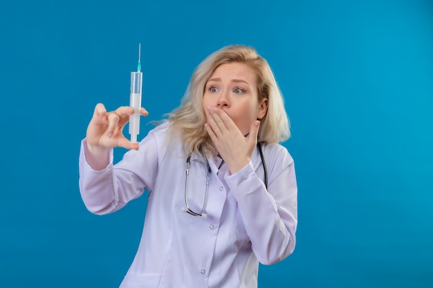 Stetoscopio da portare sorpreso della ragazza del medico in bocca coperta della siringa della tenuta dell'abito medico su fondo blu