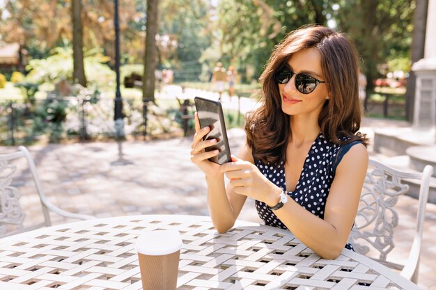 Stile bella donna con corti capelli scuri e sorriso affascinante è seduta nella caffetteria estiva alla luce del sole con il suo telefono.