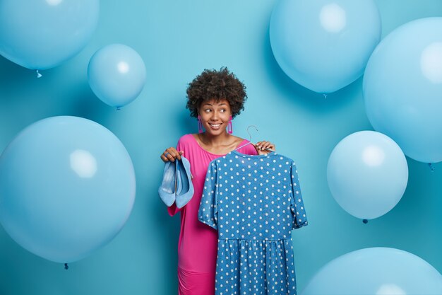 Stile, concetto di vestiti. La donna alla moda compra un vestito per un'occasione speciale, aspetta la festa, tiene un vestito a pois fantasia su grucce e scarpe blu con i tacchi alti, circondata da grandi palloncini gonfiati