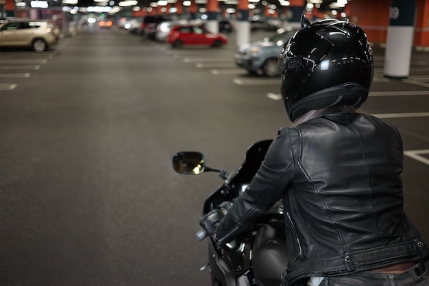 Stile di vita attivo, motociclismo, città notturna e concetto di persone. Colpo posteriore del motociclista femminile fiducioso alla moda che indossa il casco di sicurezza e giacca di pelle nera, in sella alla sua moto sul parcheggio