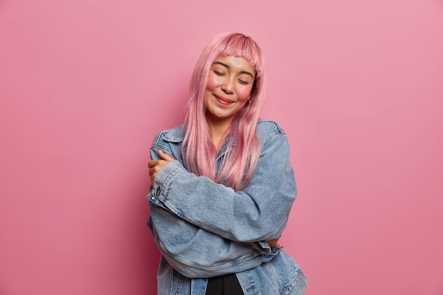 Stile di vita, concetto di emozioni umane. Felice donna tenera con lunghi capelli rosei, abbraccia il proprio corpo e ama se stessa, sta con gli occhi chiusi, indossa una giacca di jeans