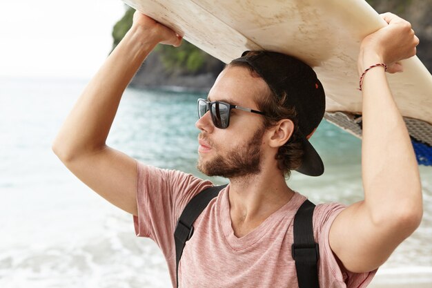 Stile di vita da surfista durante le vacanze estive. Chiuda sul ritratto di giovane atleta caucasico abbronzato attraente e bello che indossa gli occhiali da sole che escono il surf