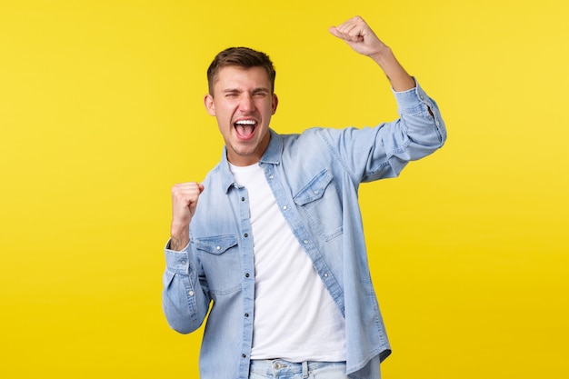 Stile di vita, emozioni delle persone e concetto di svago estivo. Entusiasta bell'uomo felice alzando le mani, cantando e gridando sì come vincente, trionfando sul premio della lotteria, sfondo giallo.