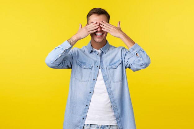 Stile di vita, emozioni delle persone e concetto di svago estivo. Sorridendo felice bell'uomo chiude gli occhi con le mani e anticipando per la sorpresa, in piedi ottimista alla festa di compleanno, sfondo giallo.