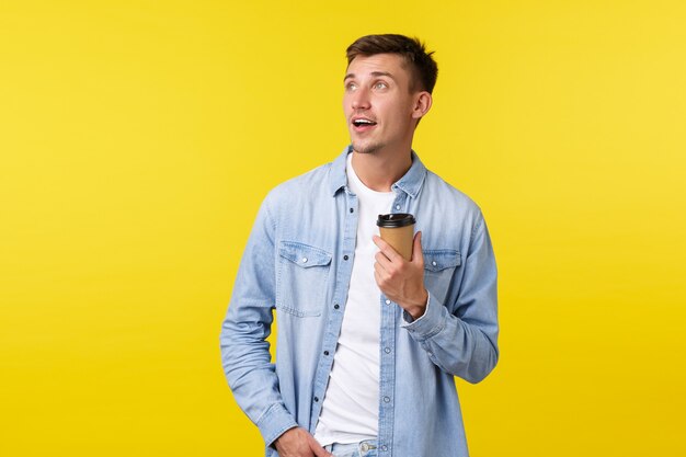 Stile di vita, emozioni delle persone e concetto di tempo libero. Il bel ragazzo biondo sognante ordina il caffè dal bar dell'ufficio durante il pranzo, guardando a sinistra premuroso mentre beve da un bicchiere di carta da asporto, sfondo giallo.