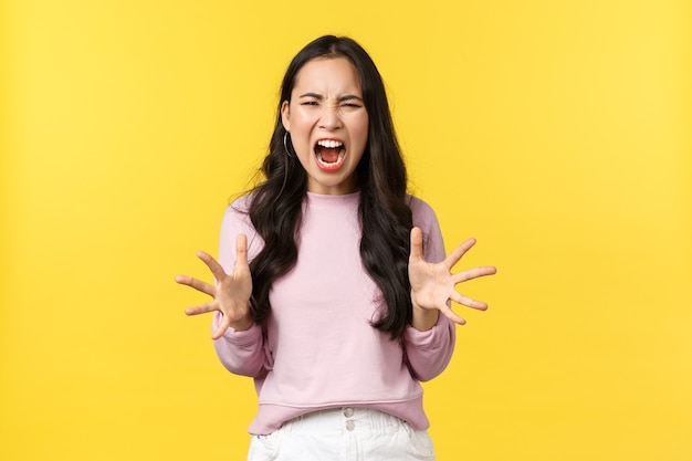 Stile di vita, emozioni e concetto di pubblicità. Ragazza coreana pazza e angosciata che perde le staffe, si sente arrabbiata e sopraffatta, urla e stringe la mano aggressiva, in piedi sfondo giallo.