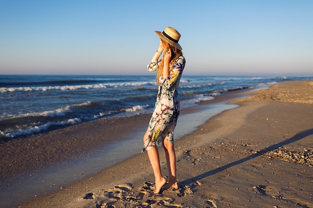 Stile di vita estate moda ritratto di bellezza donna bionda in posa sulla spiaggia solitaria, indossando bikini elegante pareo e cappello, guarda l'oceano, l'umore delle vacanze di lusso, colori dai toni luminosi.