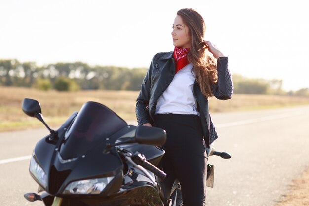 Stile di vita, estremo e concetto di persone. Colpo laterale di una giovane autista femmina piuttosto premurosa vestita in abiti alla moda, si trova vicino alla moto preferita, posa all'aperto, gode di una guida veloce