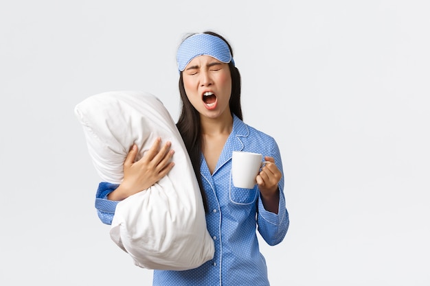 Stile di vita mattutino, colazione e concetto di persone. Ragazza con insonnia in maschera per dormire e pigiama, abbracciando il cuscino, bevendo caffè e sbadigliando, cercando di svegliarsi, sfondo bianco