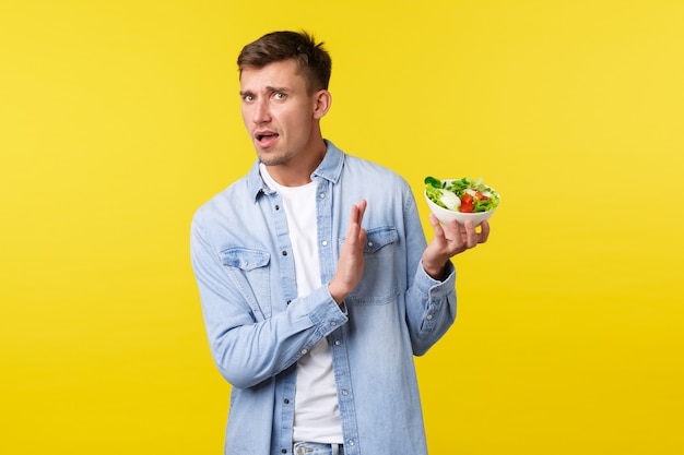 Stile di vita sano, persone e concetto di cibo. All'uomo bello allarmato e infastidito non piace mangiare questo, mostrando il gesto di rifiuto alla ciotola con insalata disgustosa, in piedi su sfondo giallo.