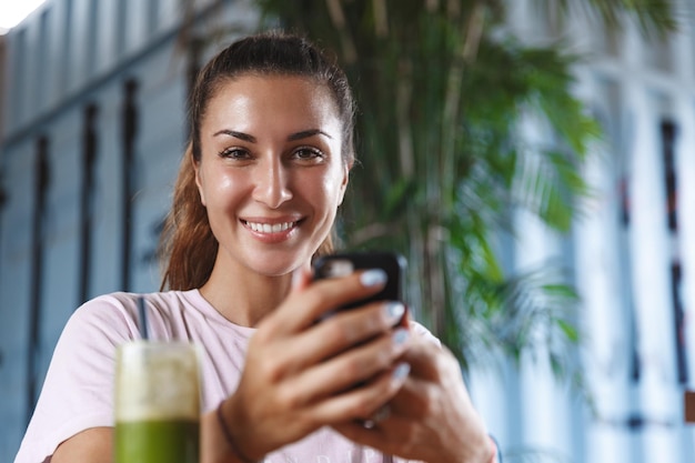 Stile di vita sano persone e concetto di svago Ritratto del primo piano della donna turistica femminile caucasica graziosa che utilizza il telefono cellulare mentre beve frullato in vacanza in viaggio verso la bellissima isola di relax