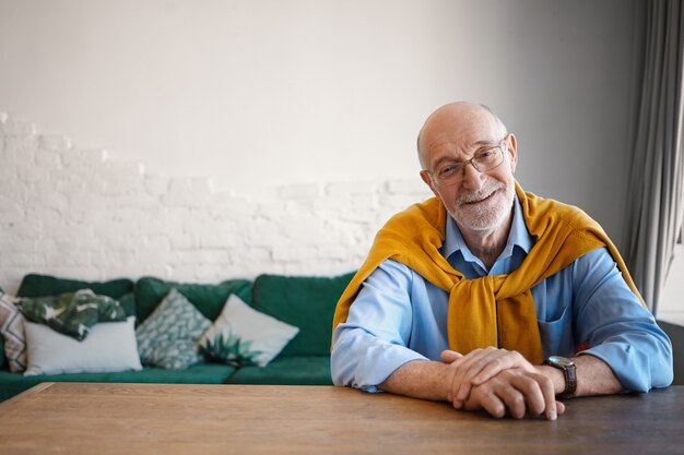 Stile, età, moda e concetto di business. Tiro al coperto di bello amichevole uomo maturo con barba grigia e testa calva lavora alla scrivania da casa, sorridente ampiamente, vestito con abiti formali