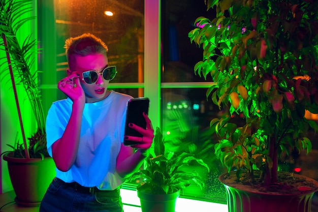 Stile. Ritratto cinematografico di donna alla moda in interni illuminati al neon. Tonica come effetti cinematografici, colori luminosi al neon. Modello caucasico utilizza lo smartphone in luci colorate al chiuso. Cultura giovanile.