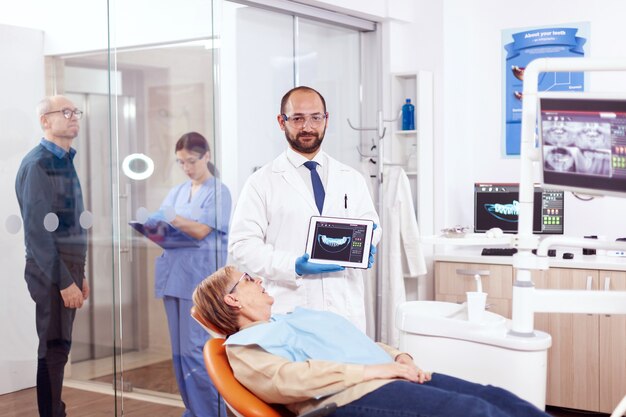 Stomatolog che tiene i raggi x della donna anziana che si siede sulla sedia arancione nell'armadietto del dentista. Addetto alla cura dei denti medici che tiene la radiografia del paziente su tablet pc vicino al paziente in piedi.