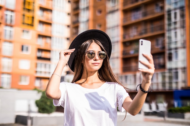 Strada all'aperto di giovane donna alla moda che fa selfie sulla strada, indossando il cappello elegante hipster.
