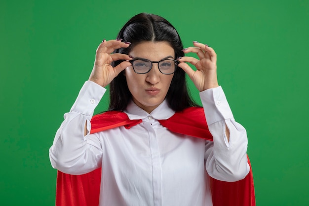 Strana giovane ragazza caucasica del supereroe che tiene le mani sugli occhiali storti isolati sulla parete verde